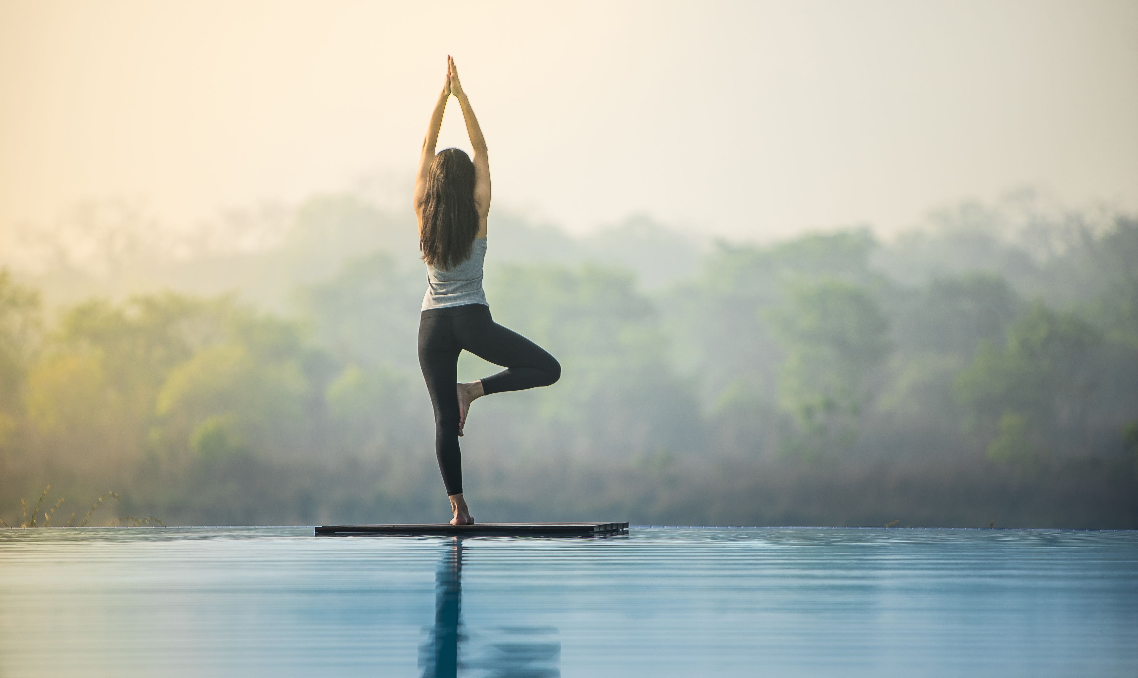 A person performing a yoga pose on a floating platform in a body of water with a forested background.