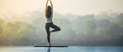 A person performing a yoga pose on a floating platform in a body of water with a forested background.