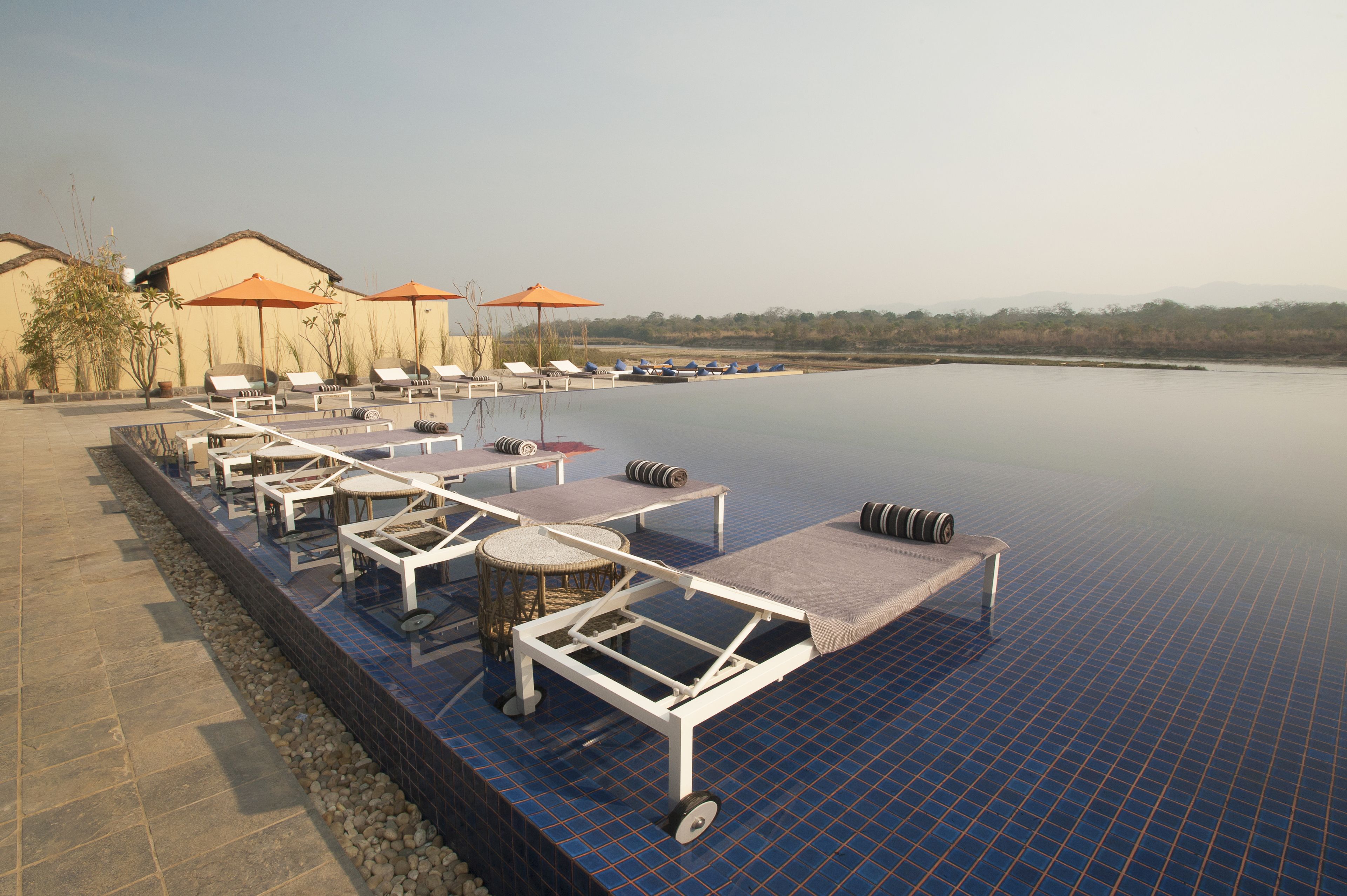 An infinity pool with lounge chairs and umbrellas, a tiled deck, and a view of distant hills.