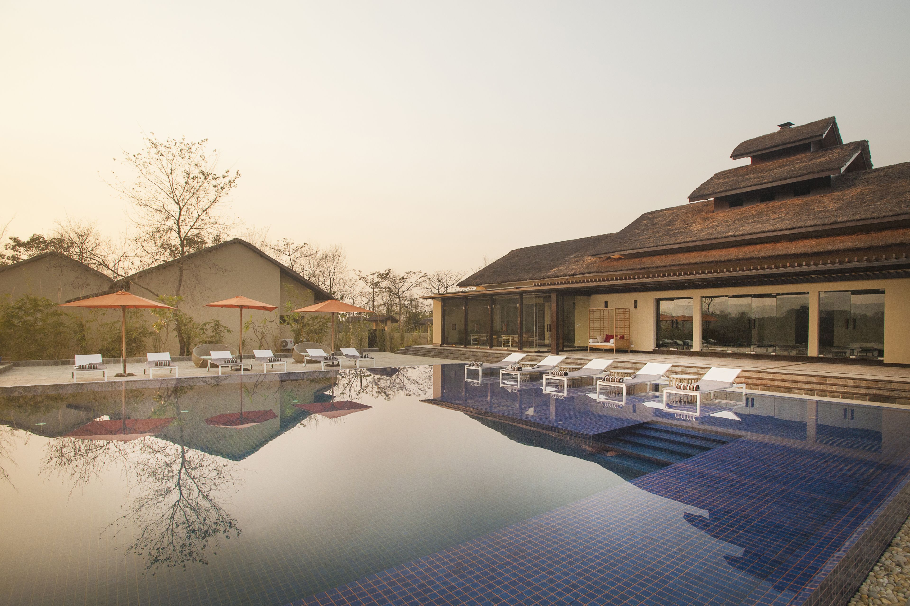 A swimming pool with lounge chairs and umbrellas, surrounded by buildings with large windows and a tiled roof.