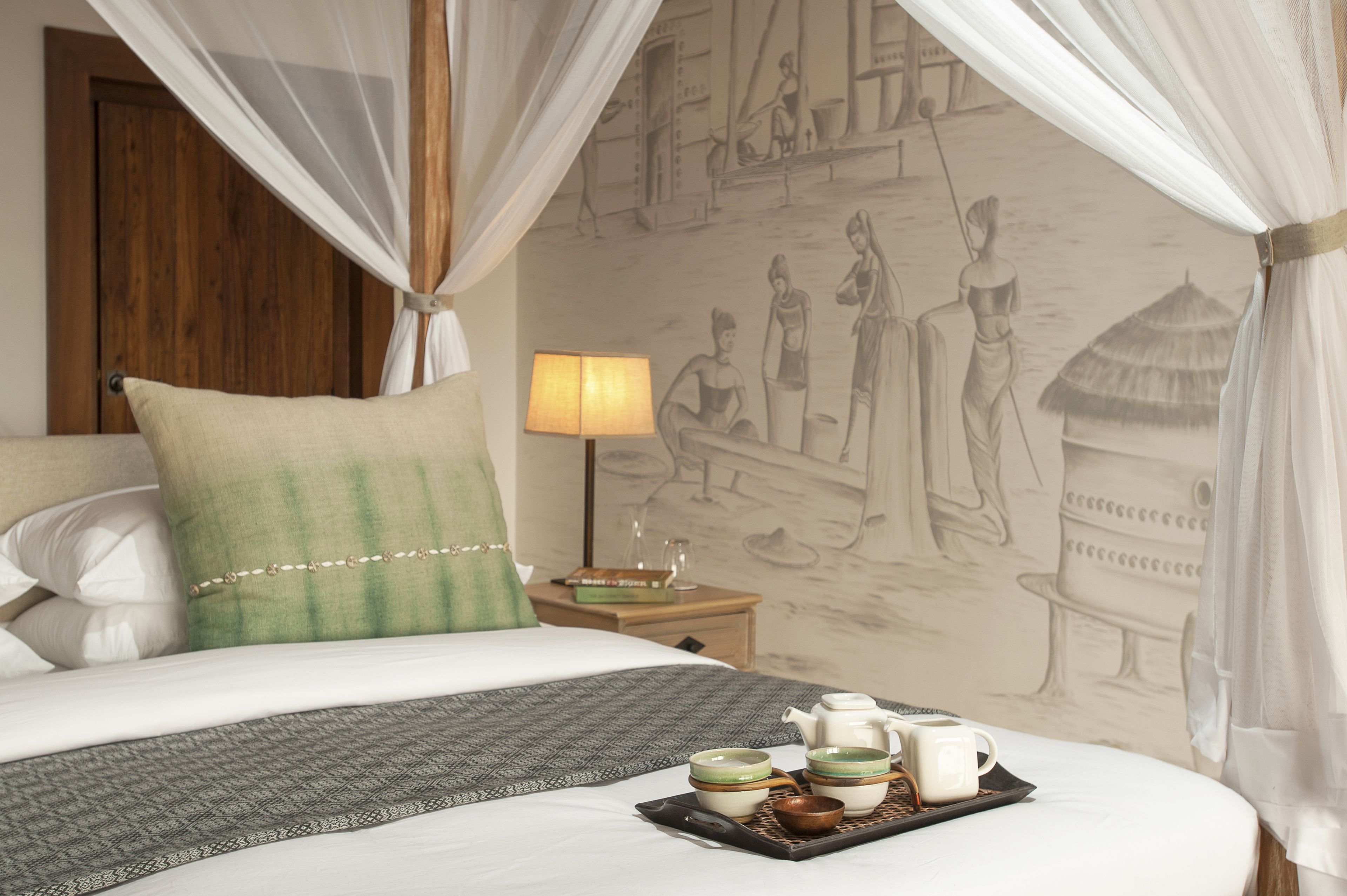 Habitación de hotel con cama, mesita de noche con lámpara y bandeja con té y tazas.