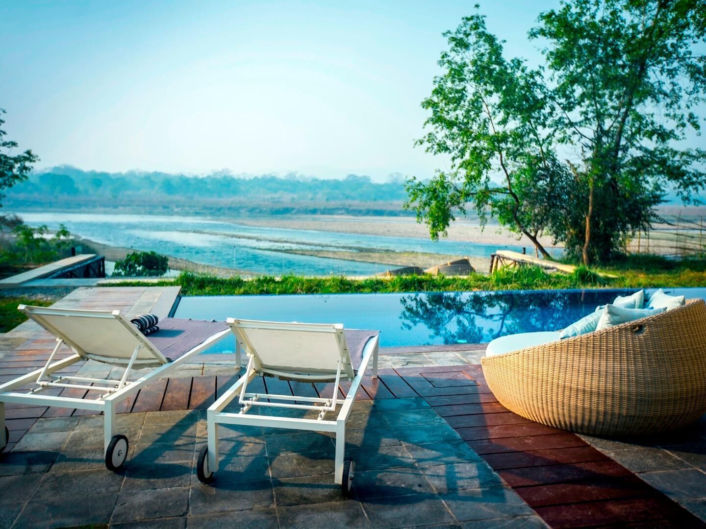 A poolside area with lounge chairs and a wicker ottoman, overlooking a body of water and greenery.
