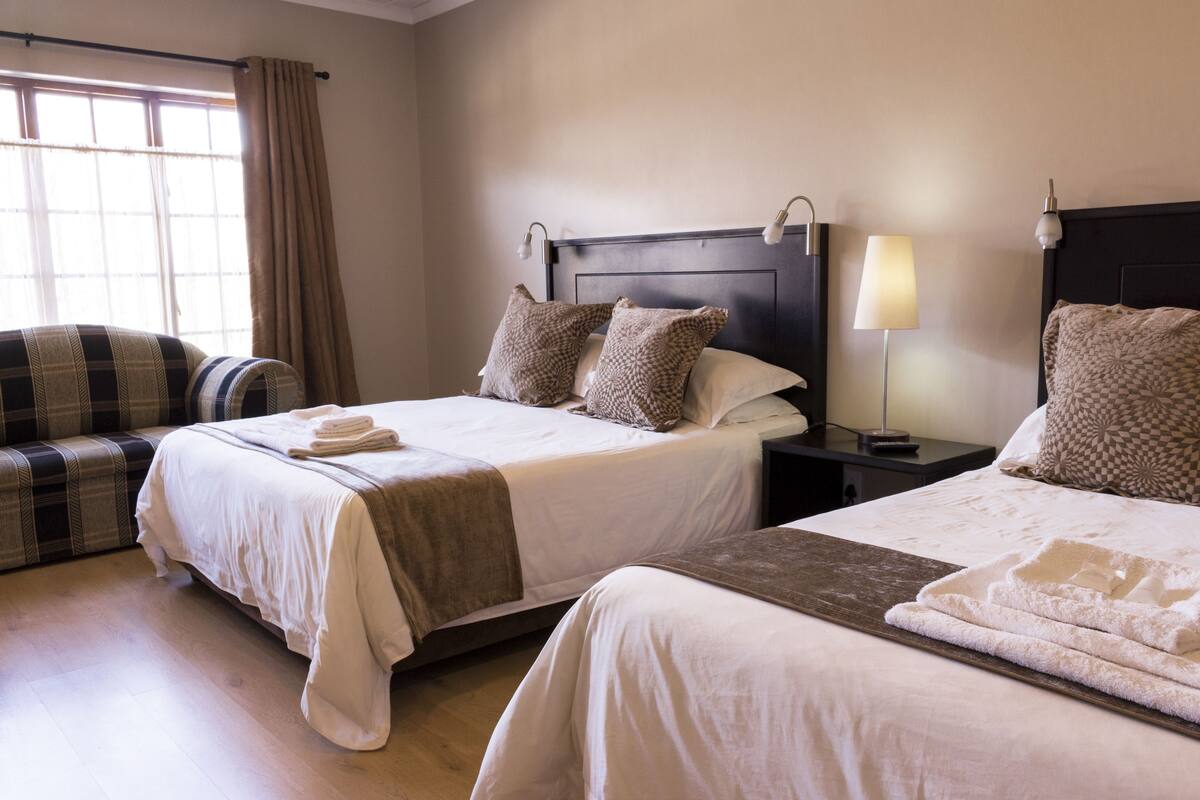 Une chambre d’hôtel avec deux lits, un canapé, une table de chevet avec une lampe et une fenêtre avec des rideaux.