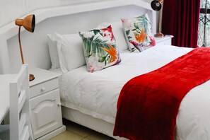 A neatly made bed with white bedding and a red throw blanket, accompanied by two decorative pillows with a tropical print.