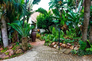 A garden pathway leading to a building with a tiled roof, surrounded by lush tropical plants and a large decorative vase.