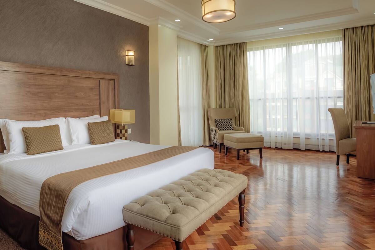 A hotel room with a large bed, two bedside lamps, a wooden headboard, a bench, a chair, a desk, and a large window with curtains.