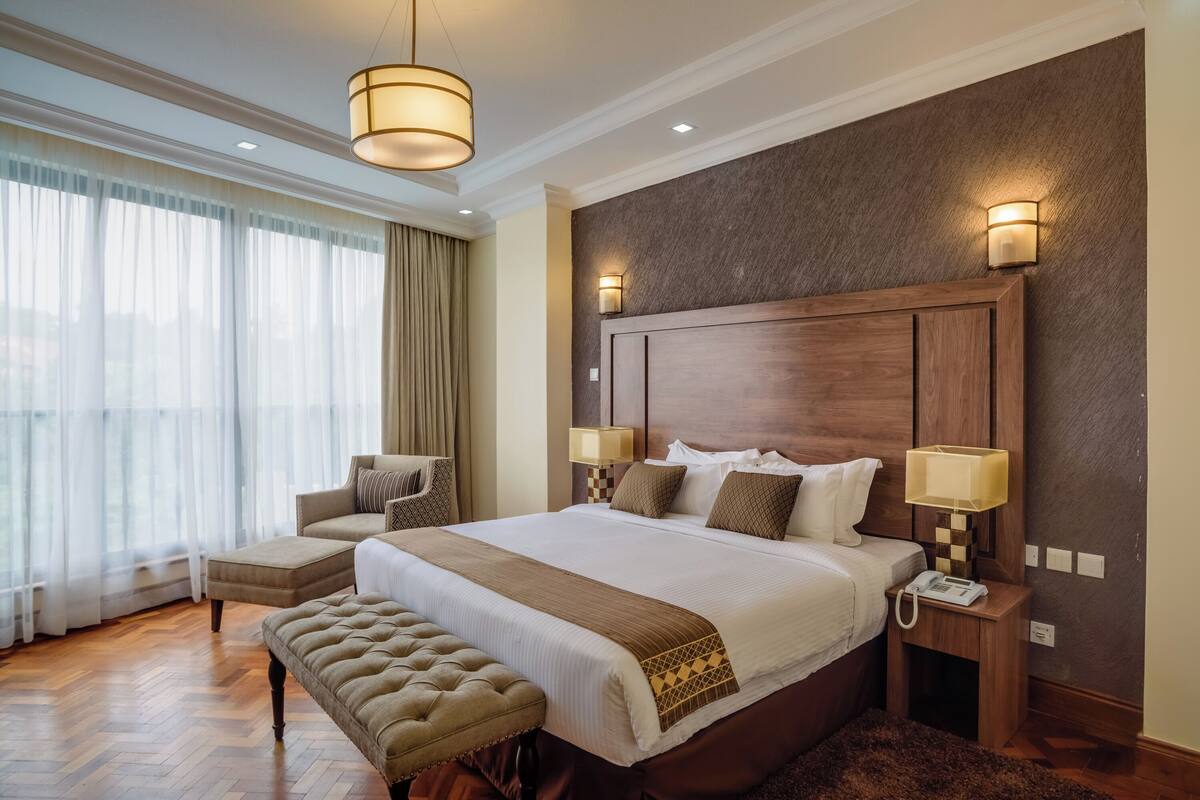 A spacious bedroom with a large bed, two bedside tables with lamps, a wooden headboard, and a view of the outdoors through sheer curtains.