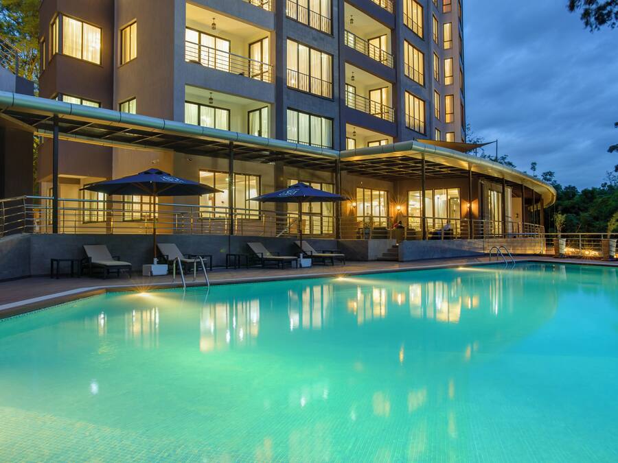 Una piscina con sillones de descanso y sombrillas, una zona cubierta con asientos, y un edificio de varios pisos con balcones y ventanas.