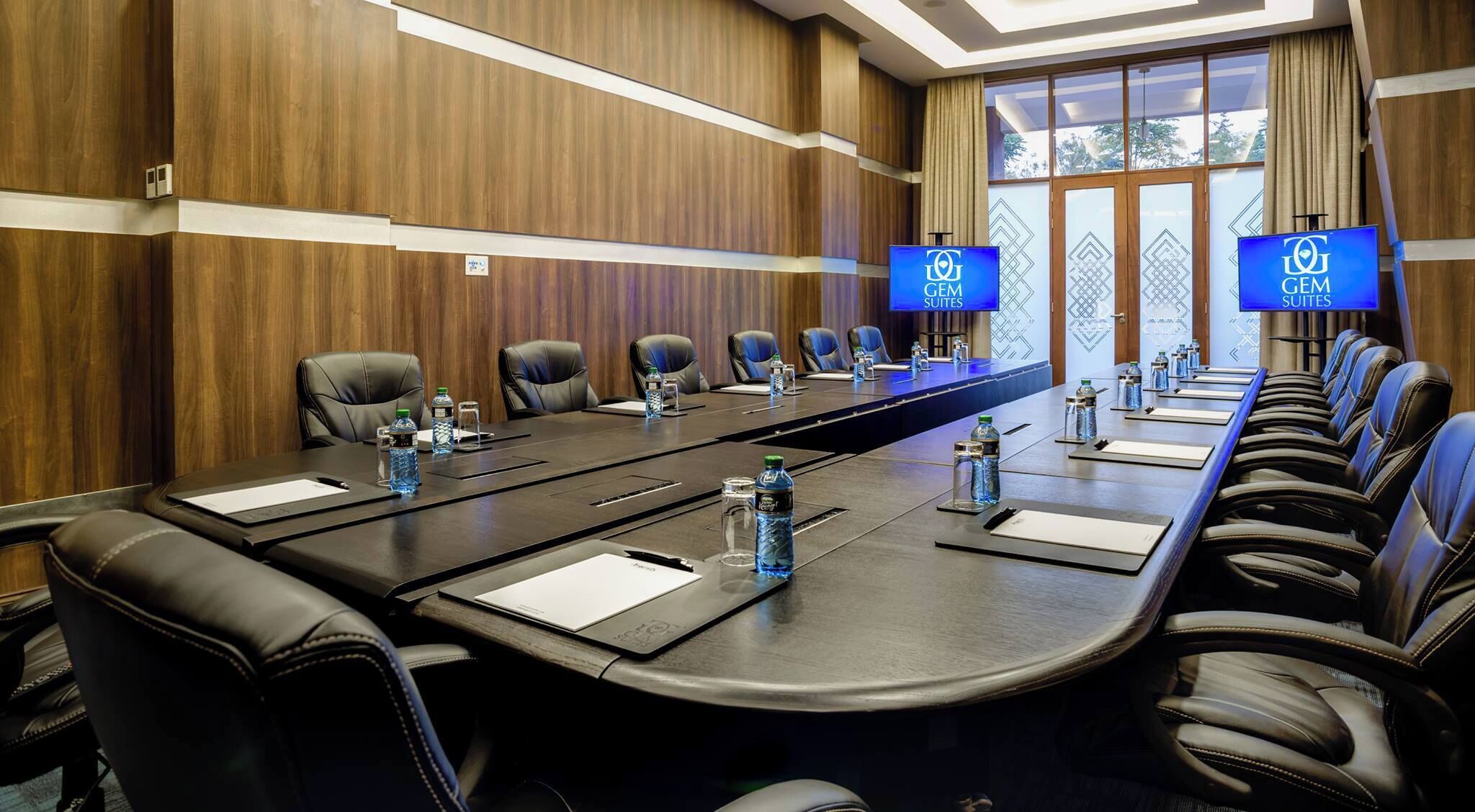 Une salle de conférence équipée d’une longue table, de nombreuses chaises, de bouteilles d’eau et de deux écrans affichant le texte « GEM SUITES ».