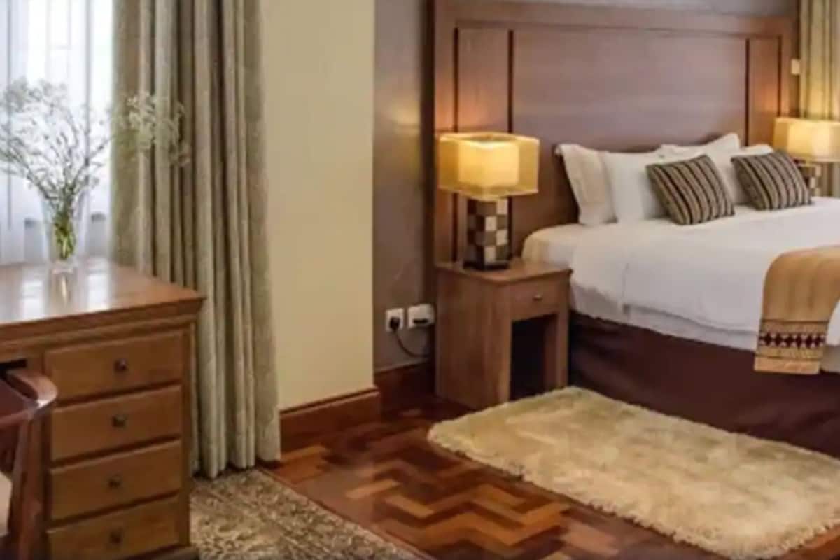 A hotel room with a wooden desk, a bed with a white comforter and striped pillows, a nightstand with a lamp, and a small rug.