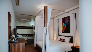 A modern hotel room with a four-poster bed, a bedside table, a nightstand, a desk with a chair, a mirror, and a bathroom visible in the background. - Hotel Select (Sibiu)