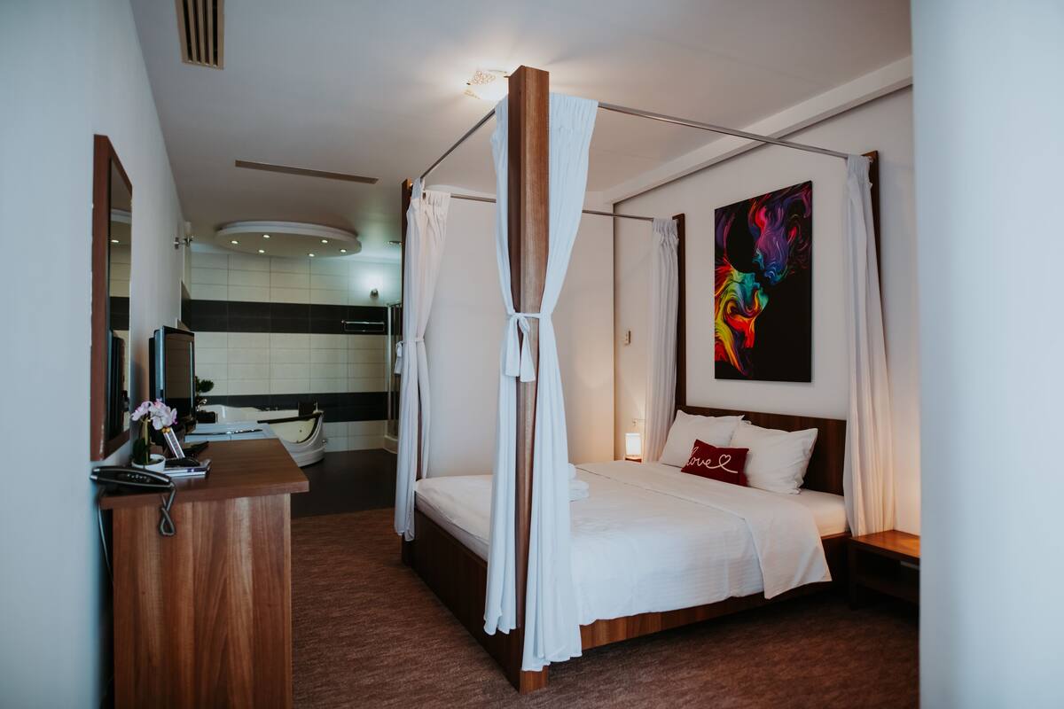 Une chambre d’hôtel moderne avec un lit à baldaquin, une table de chevet, une petite table, un bureau avec une chaise, un miroir et une salle de bain visible au fond.