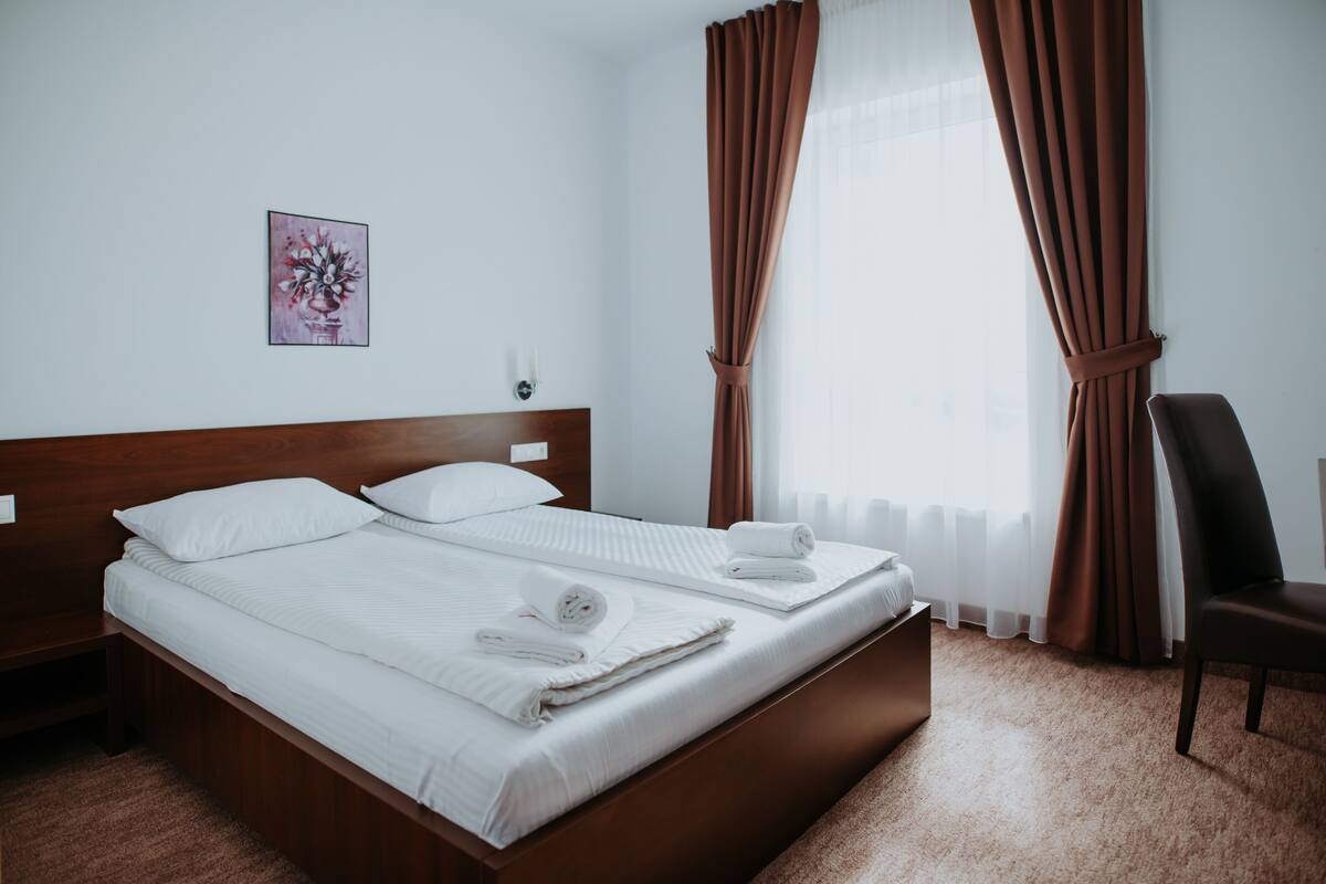 Un lit bien fait, avec des draps blancs et des serviettes pliées, une tête de lit en bois, une seule lampe fixée au mur et un tableau encadré accroché au mur.