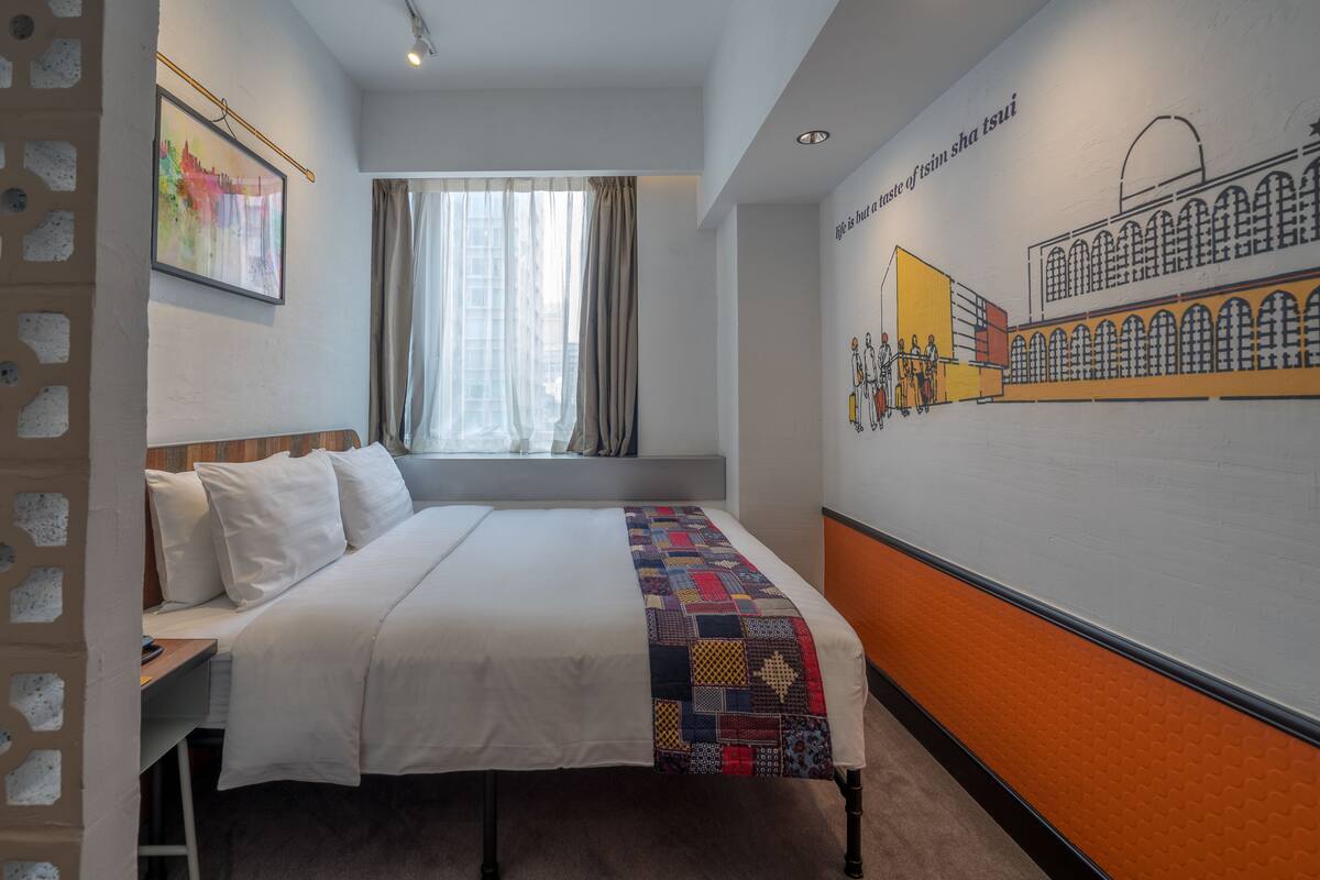 A hotel room with a bed, a window with curtains, a wall mural, and a small seating area with a bench.