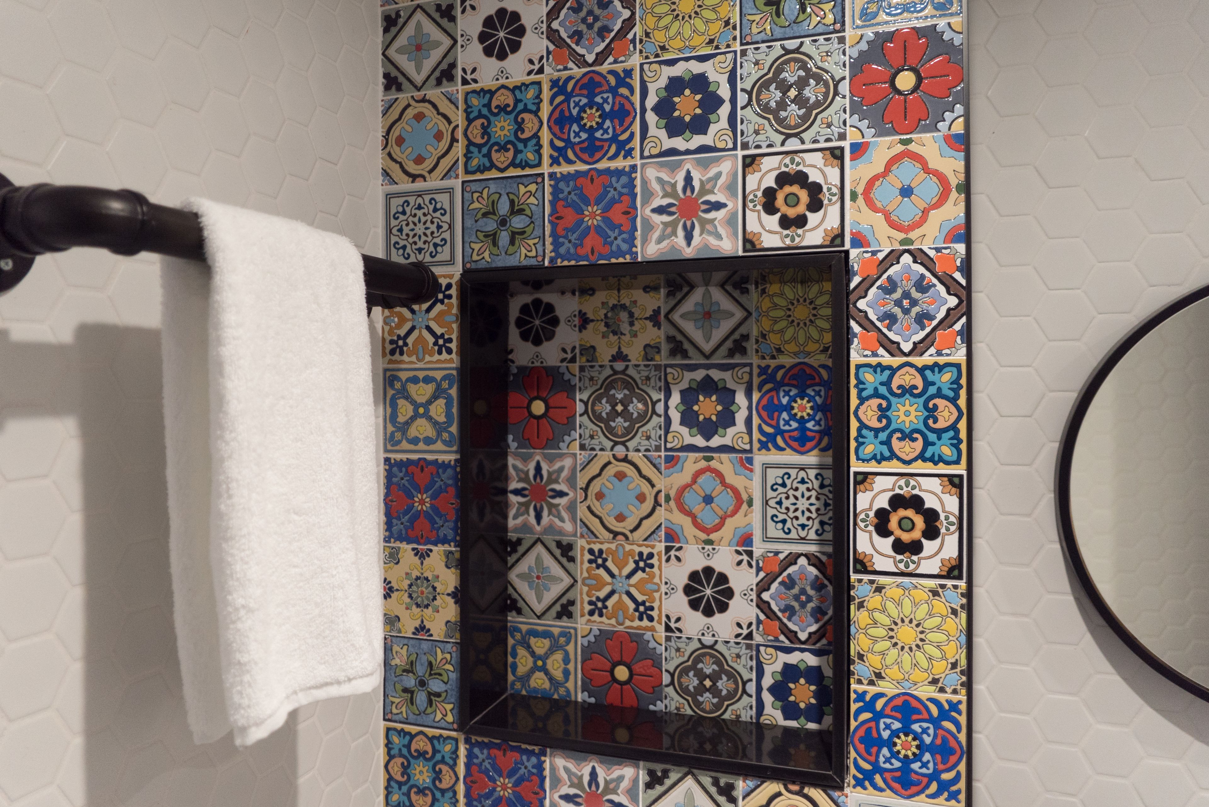 Une salle de bain avec un mur recouvert de carreaux aux motifs colorés variés, une serviette suspendue à un porte-serviettes et un miroir rond.