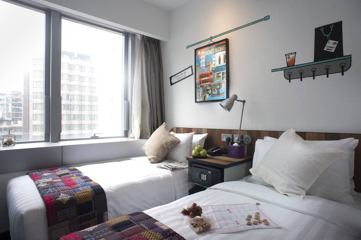 A hotel room with two beds, a window with curtains, a cityscape view, a framed picture on the wall, and a bedside table with a lamp and a glass of water.