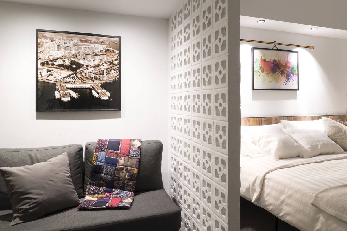 A modern hotel room with a grey sofa, a colorful quilt, a cityscape painting, a bed with white linens, and a patterned wall.
