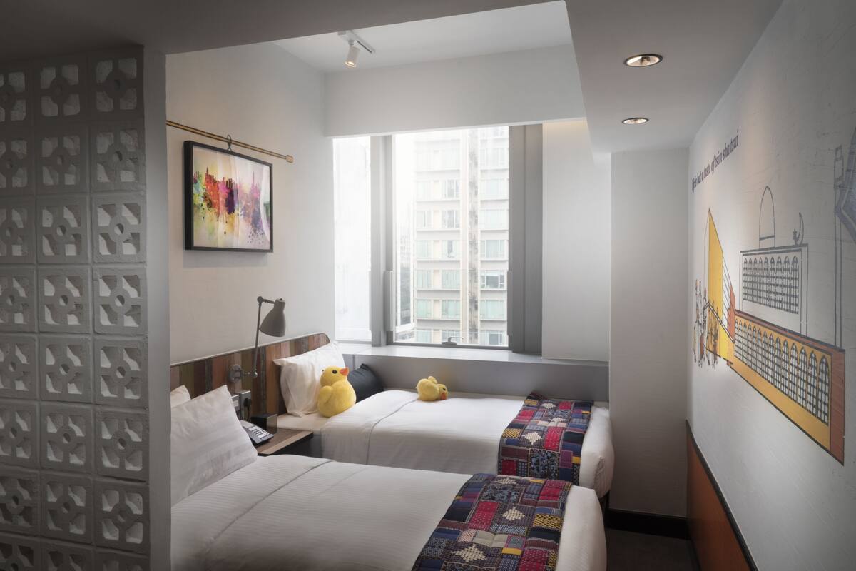 A hotel room with two beds, a window with a view of buildings, a wall with a cityscape mural, and a decorative partition.