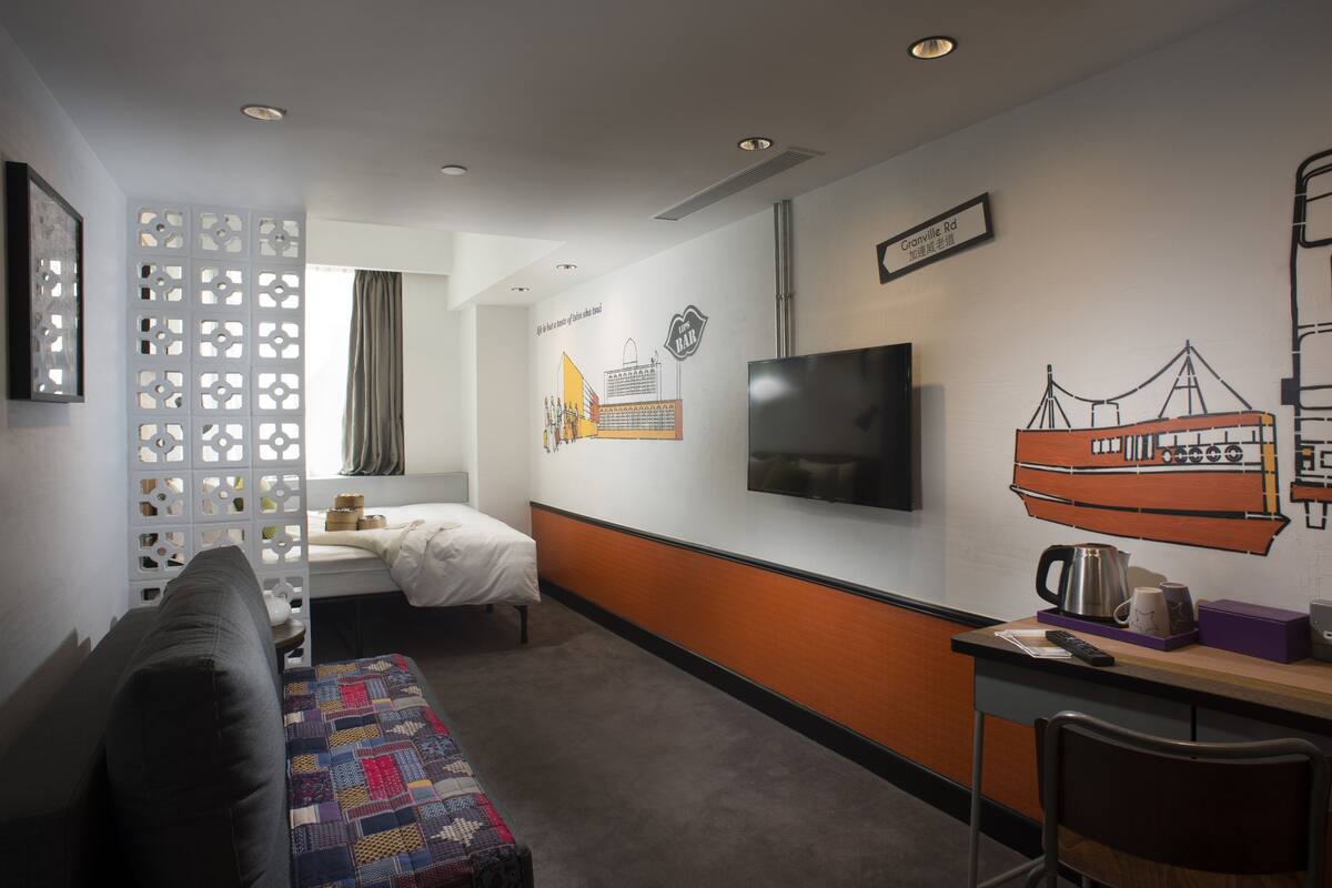 A hotel room with a bed, a sofa, a desk with a chair, a TV, and a wall with artwork.