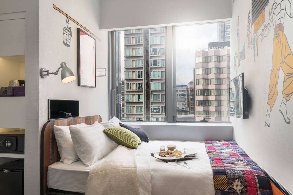A modern bedroom with a bed, a window with a city view, a bedside table with a lamp, and a wall-mounted TV.