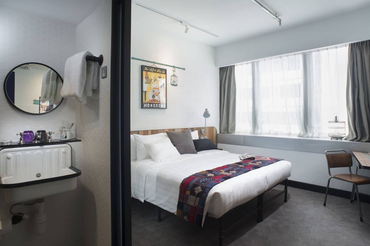 A hotel room with a bed, bedside table, chair, window with curtains, radiator, sink, mirror, and a poster on the wall.