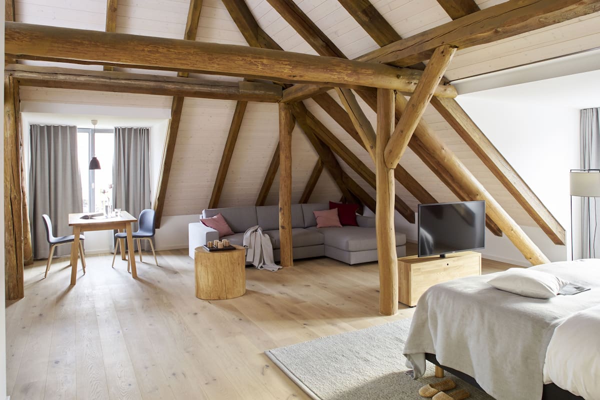 Un amplio dormitorio en el ático con vigas de madera a la vista, una cama grande, un televisor y un área de descanso con sofá y sillones.