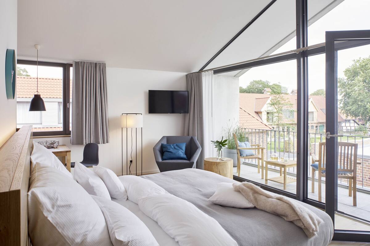 Una habitación de hotel moderna con una cama grande, un escritorio, una silla, un televisor y un balcón con mobiliario exterior.