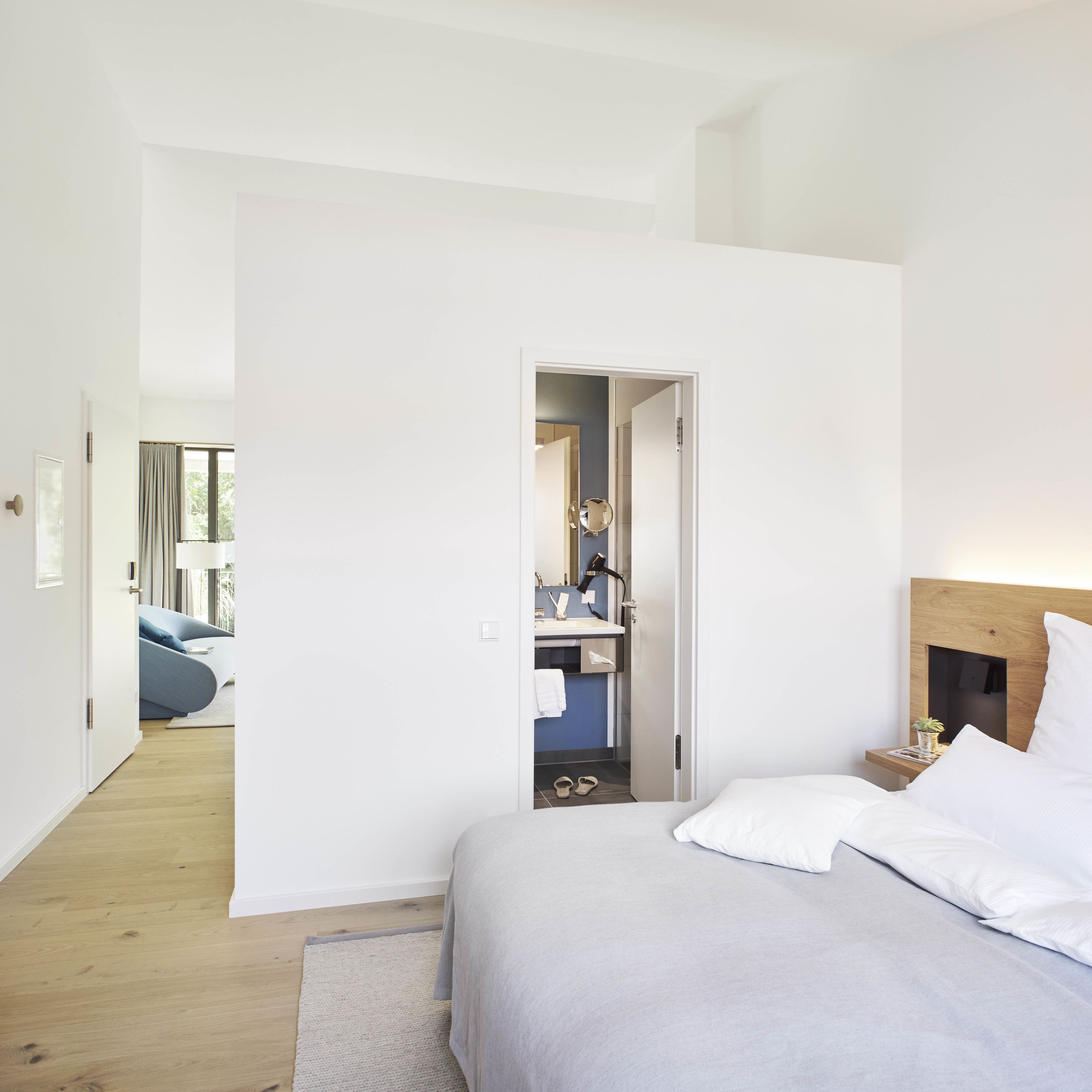 A modern bedroom with a large bed, a bedside table with a lamp, and a view into a bathroom with a mirror and towel rack.