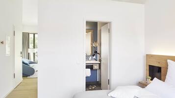 A modern bedroom with a large bed, a bedside table with a lamp, and a view into a bathroom with a mirror and towel rack.