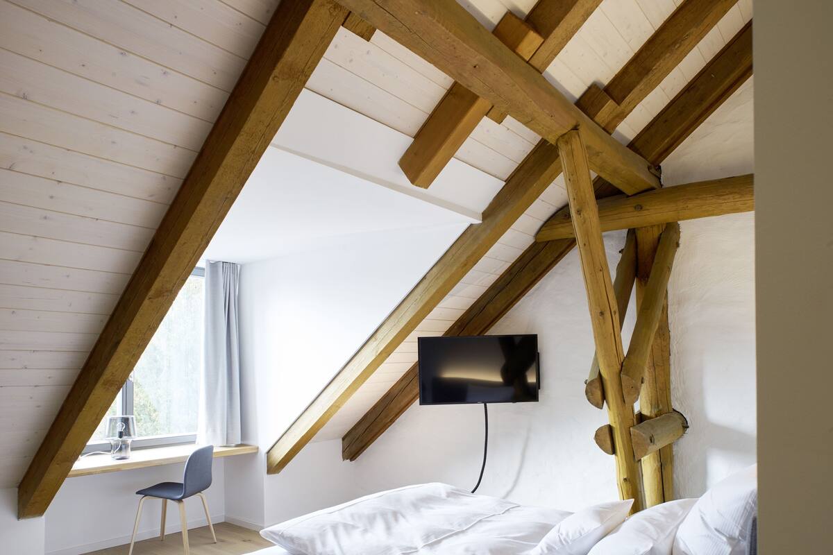Un dormitorio con techo inclinado, una cama, una silla y un televisor instalado en la pared.