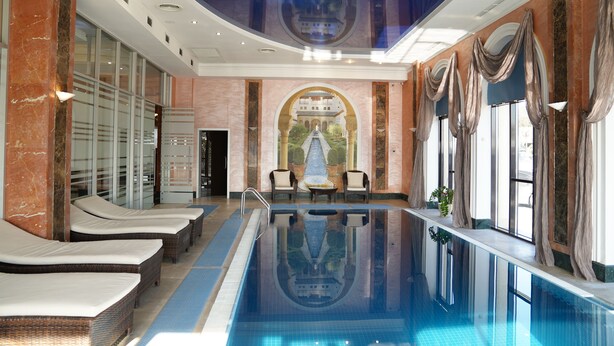 Uma grande piscina interior com espreguiçadeiras, um mural e vista para um edifício através de um arco.