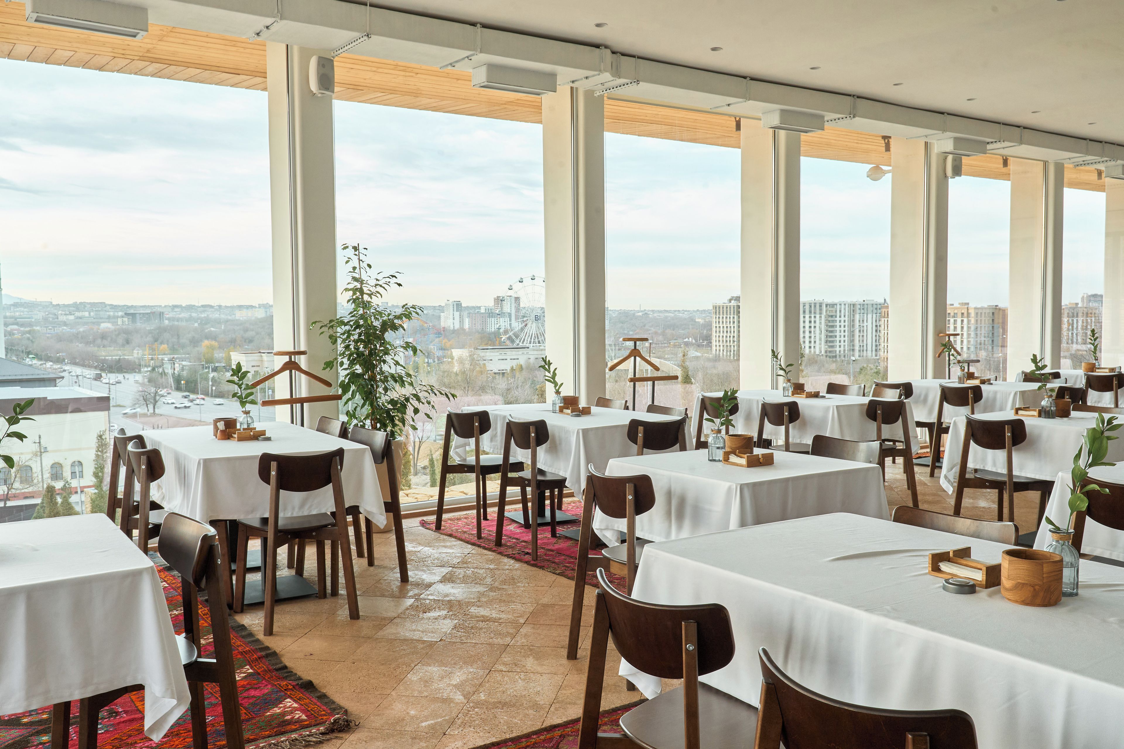 A restaurant with multiple tables and chairs, large windows offering a city view, and a red carpet leading to the entrance.