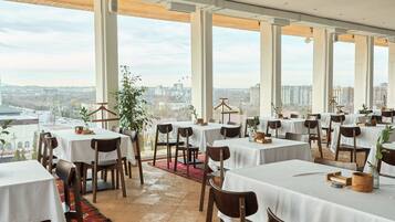 A restaurant with multiple tables and chairs, large windows offering a city view, and a red carpet leading to the entrance.