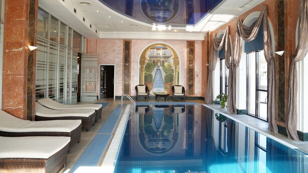 A large indoor swimming pool with lounge chairs, a mural, and a view of a building through an archway. - Kainar Hotel (Shymkent)