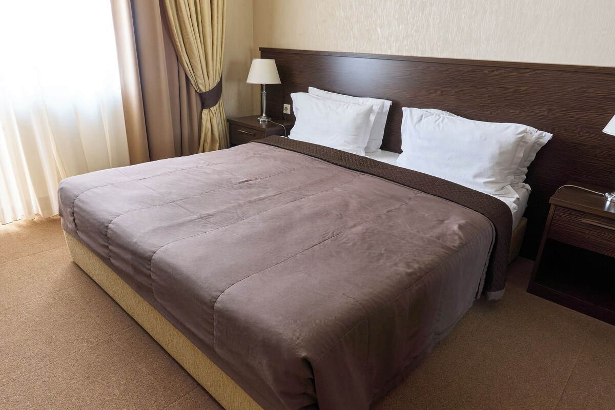 A hotel room with a large bed, a nightstand with a lamp, and a window with curtains.