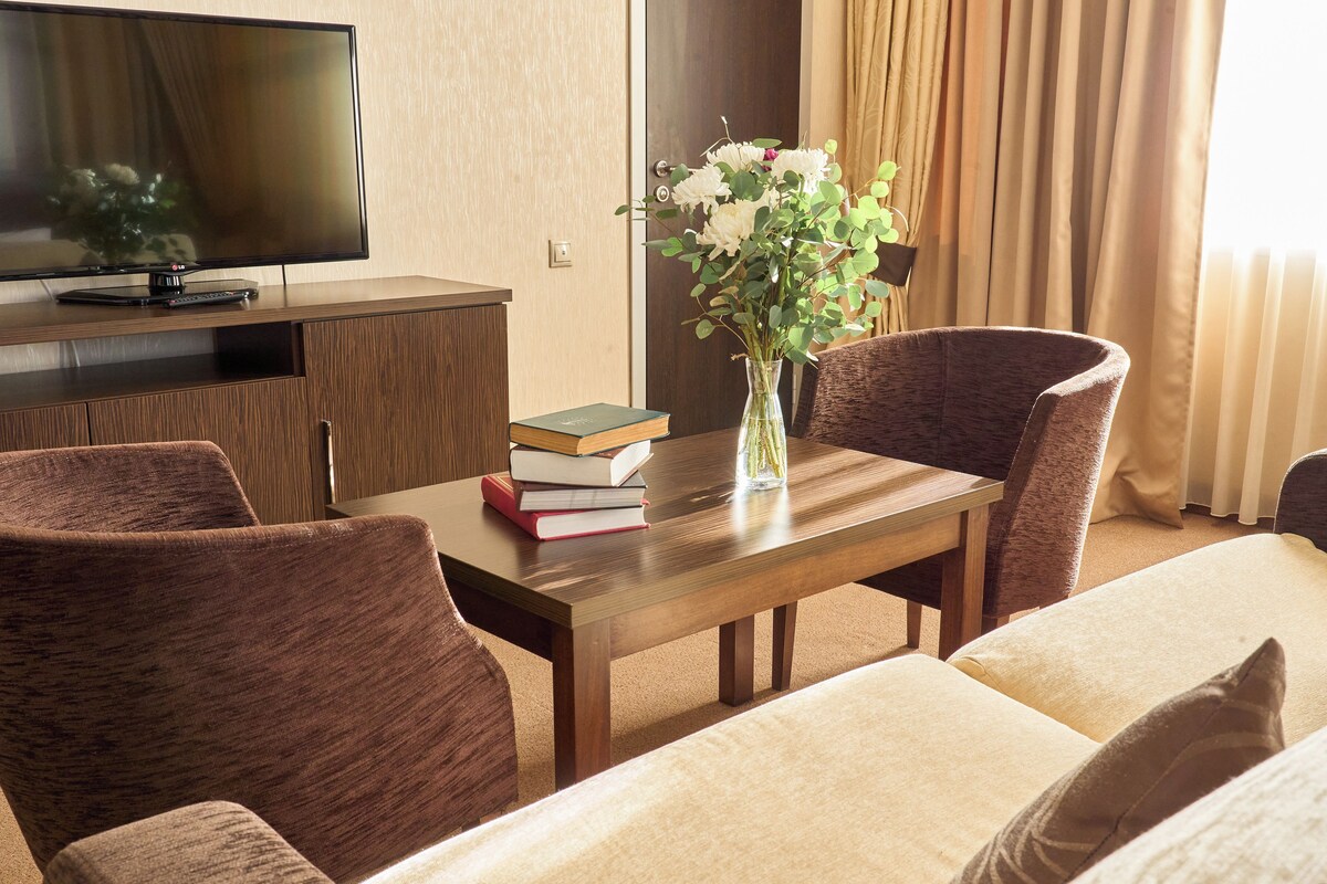 A hotel room with a wooden TV stand, a flat-screen TV, a table with a vase of flowers, and two brown upholstered chairs.