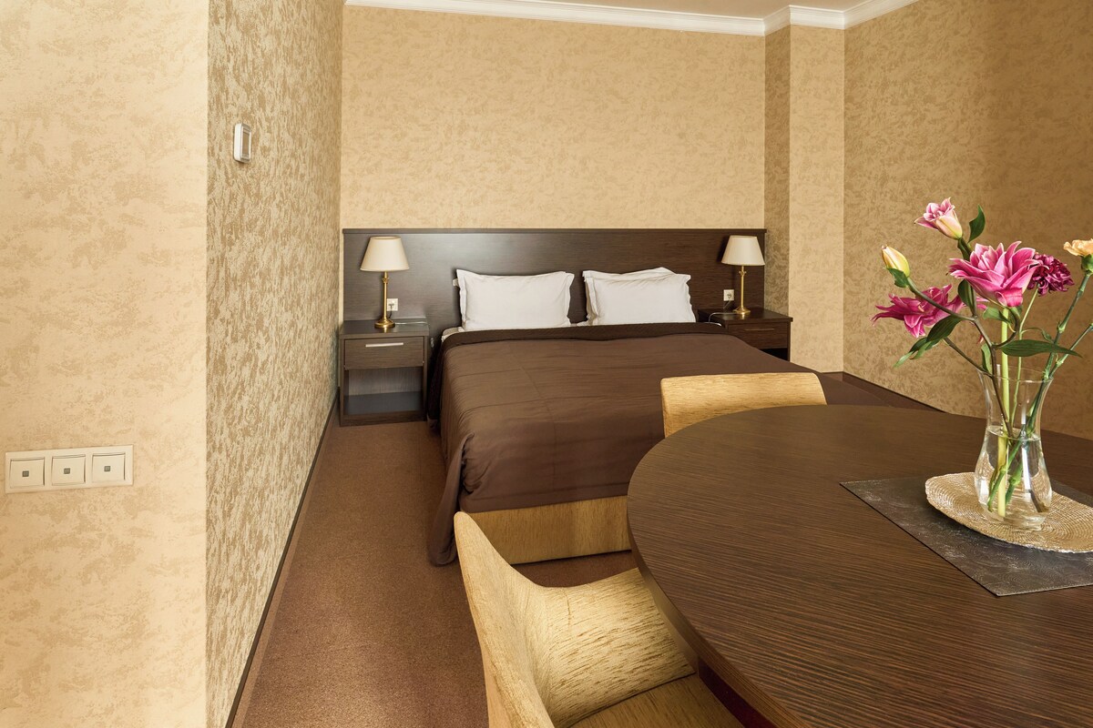 A hotel room with a bed, bedside tables, a table with chairs, and a vase with flowers.