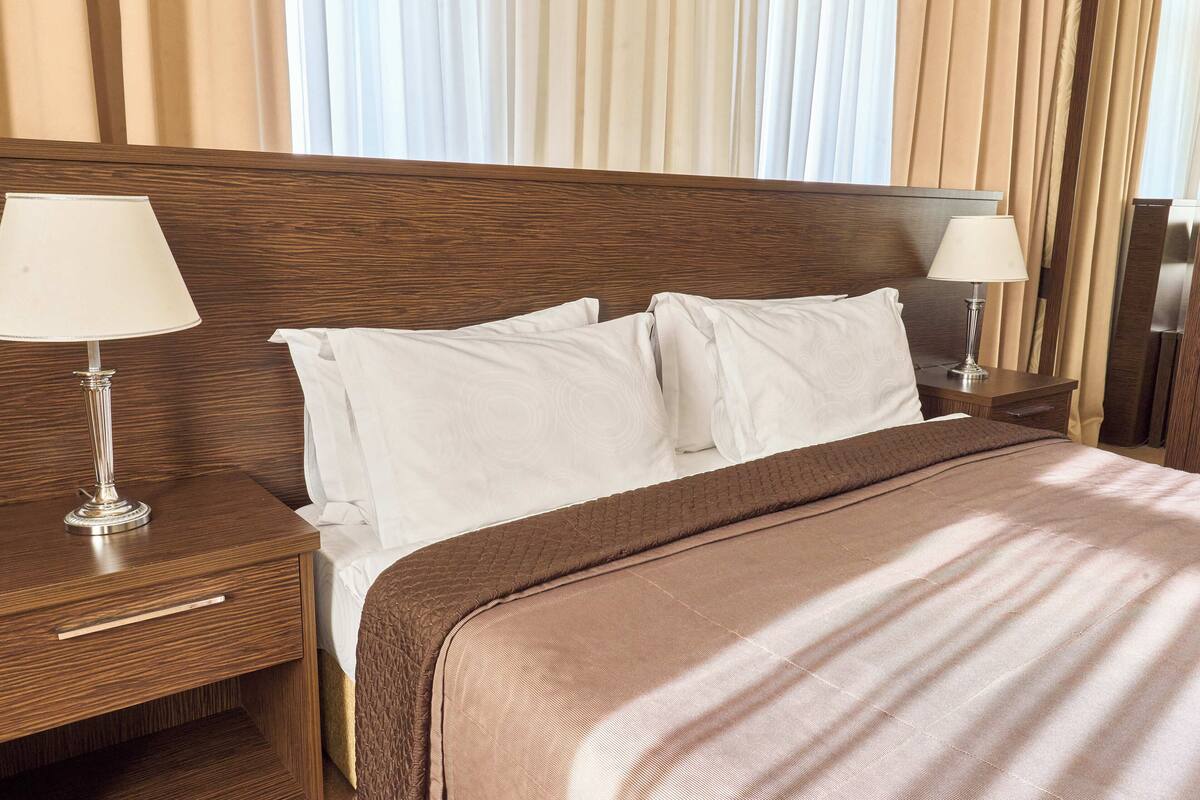 A hotel room with a wooden headboard, two bedside tables with lamps, a large bed with white linens, and a view of the curtains.