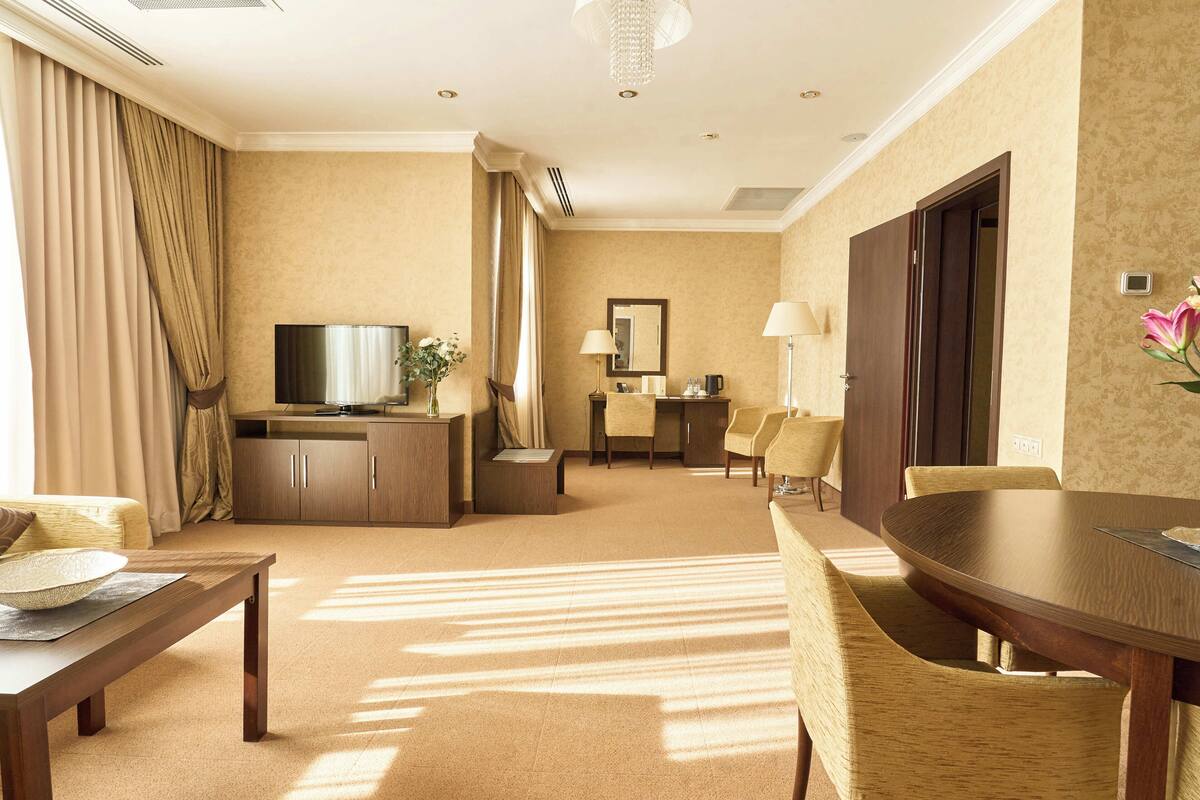 A hotel room with a dining area, a television, a desk, and a large window with curtains.