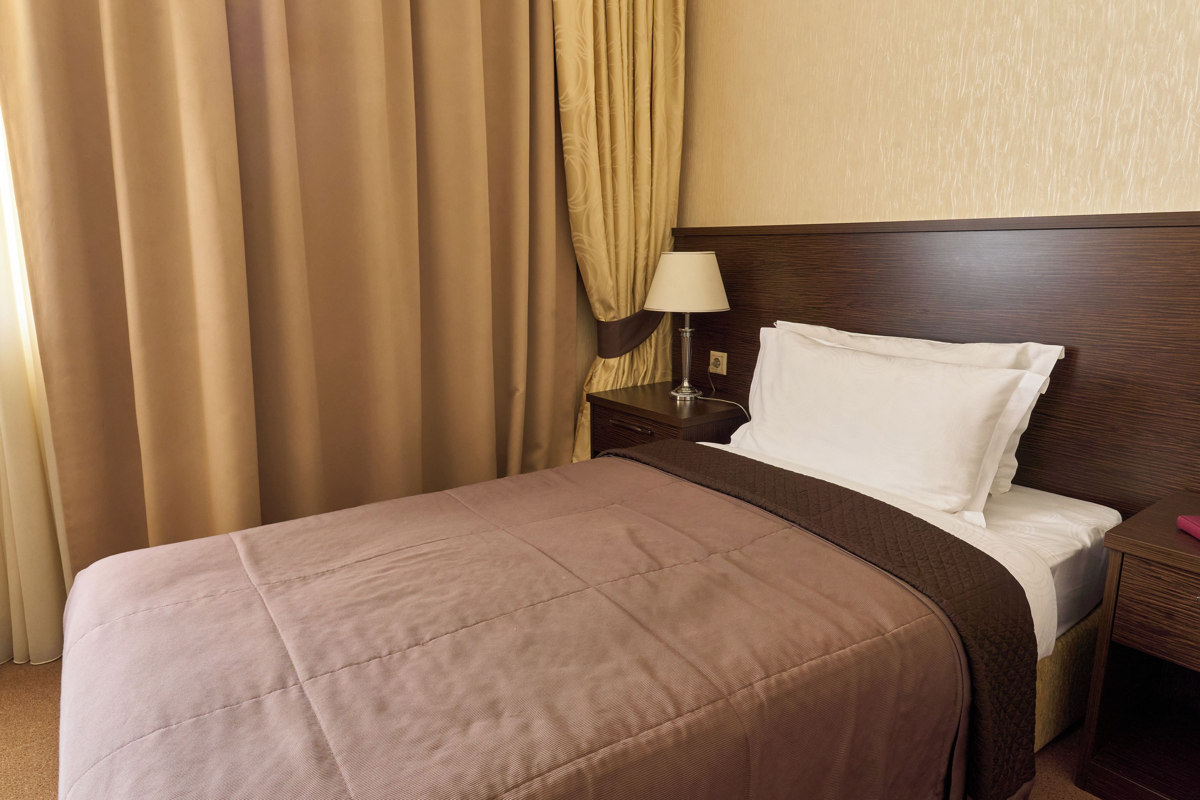 Una cama bien hecha con cubrecama marrón y almohadas blancas, una mesita de noche de madera con una lámpara y cortinas color beige.