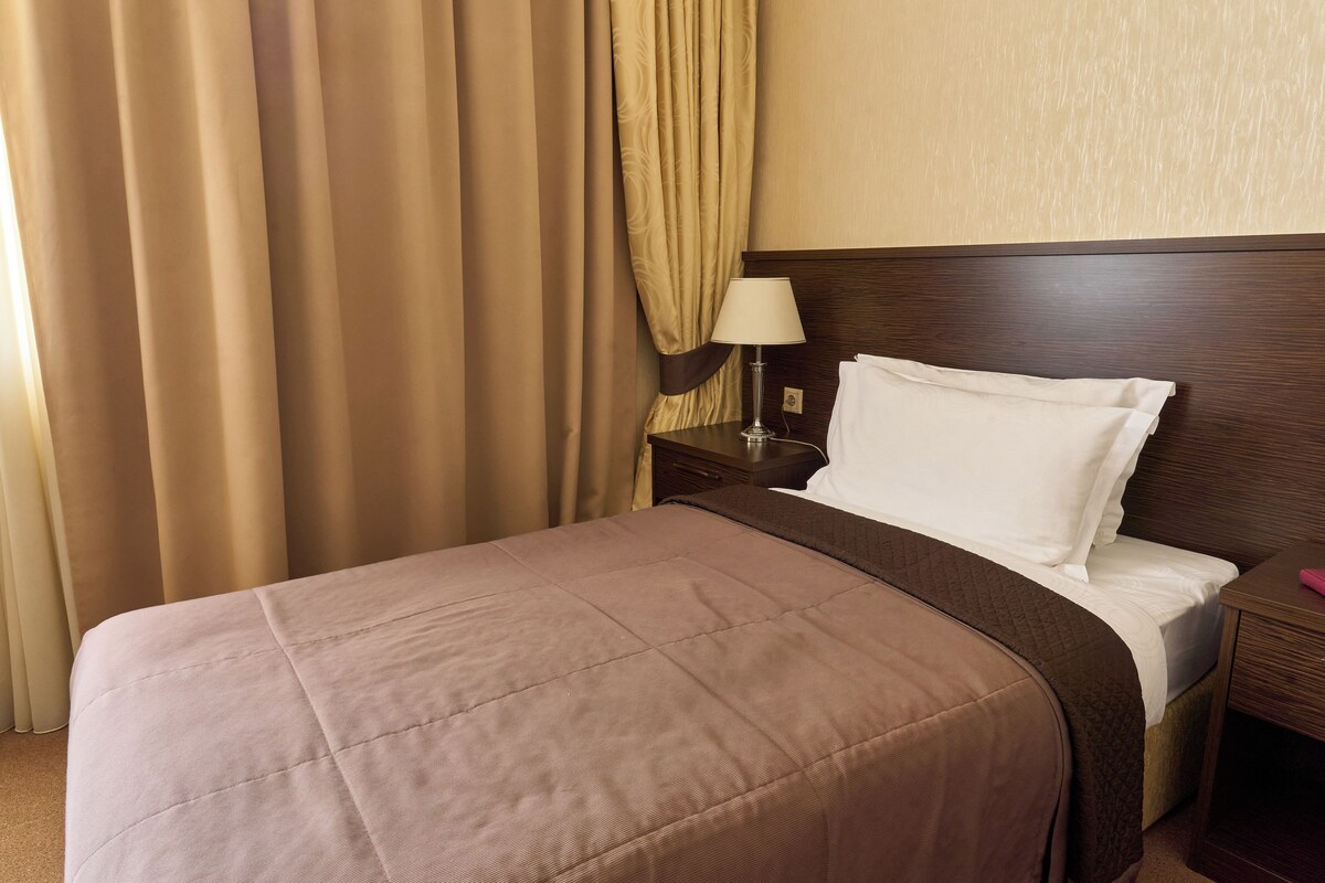 A neatly made bed with a brown bedspread and white pillows, a wooden nightstand with a lamp, and beige curtains.
