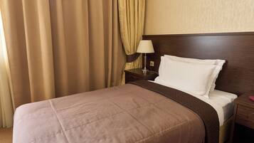 Una cama bien hecha con cubrecama marrón y almohadas blancas, una mesita de noche de madera con una lámpara y cortinas color beige.