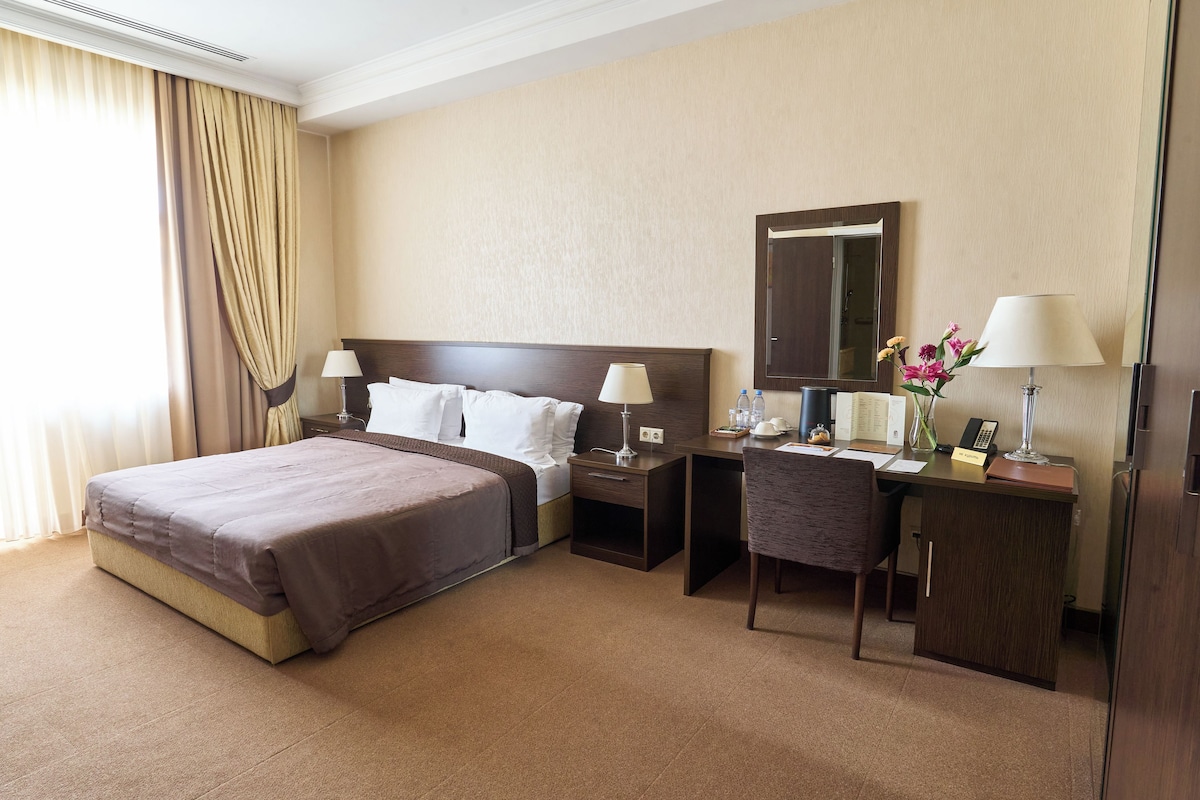 A hotel room with a large bed, a desk with a chair, a nightstand, a mirror, and a lamp.