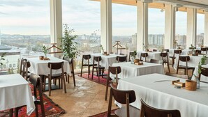 A restaurant with multiple tables and chairs, large windows offering a city view, and a red carpet leading to the entrance. - Kainar Hotel (Shymkent)