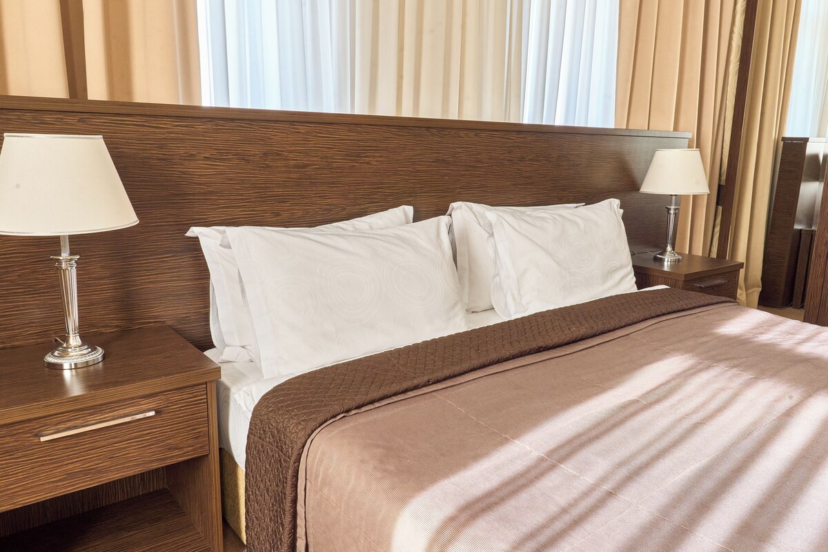 A hotel room with a wooden headboard, two bedside tables with lamps, a large bed with white linens, and a view of the curtains.