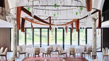 A spacious dining area with multiple tables and chairs, large windows offering a view of the outdoors, and a ceiling with hanging lights and decorative elements.