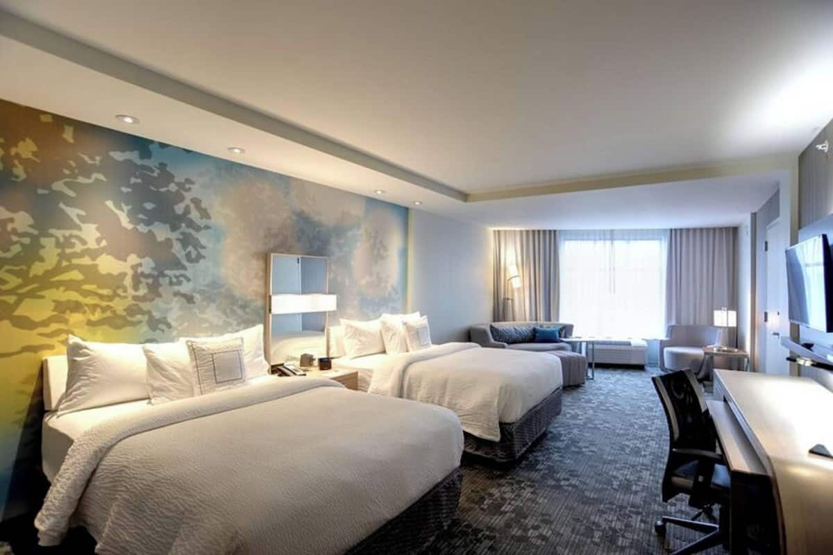 A hotel room with two beds, a desk, a chair, a TV, and a large window with curtains.
