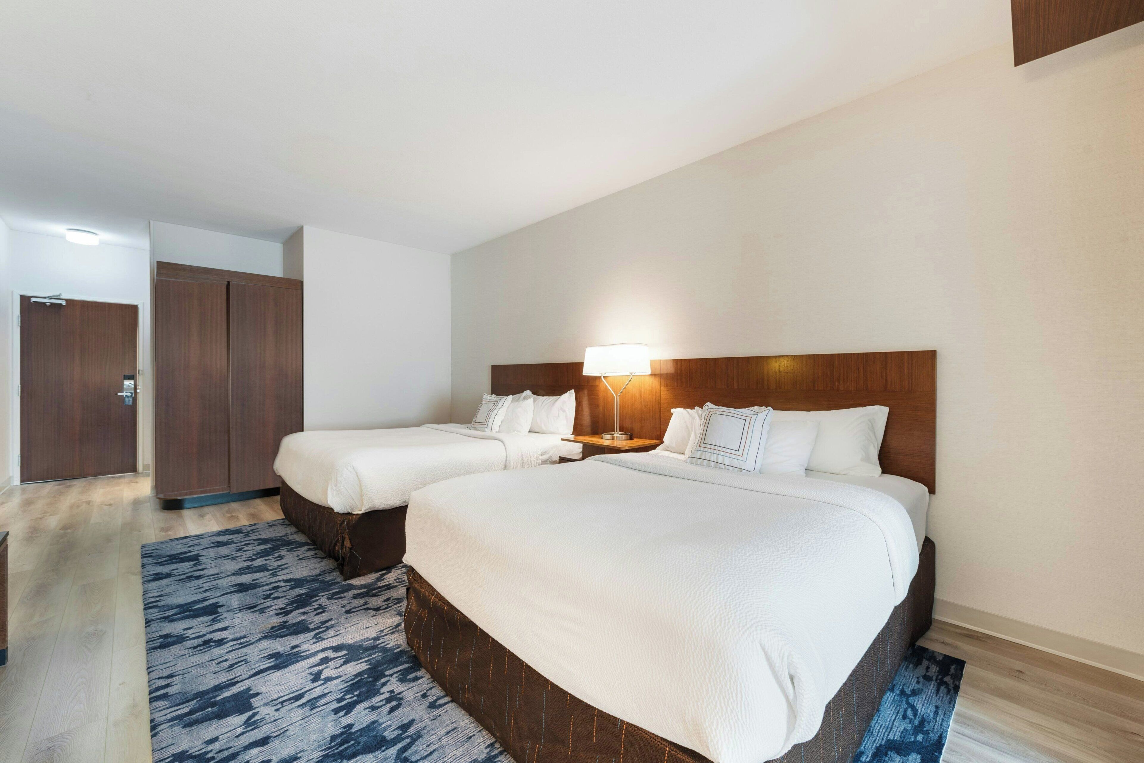 A hotel room with two beds, a wooden headboard, a nightstand with a lamp, and a patterned rug.