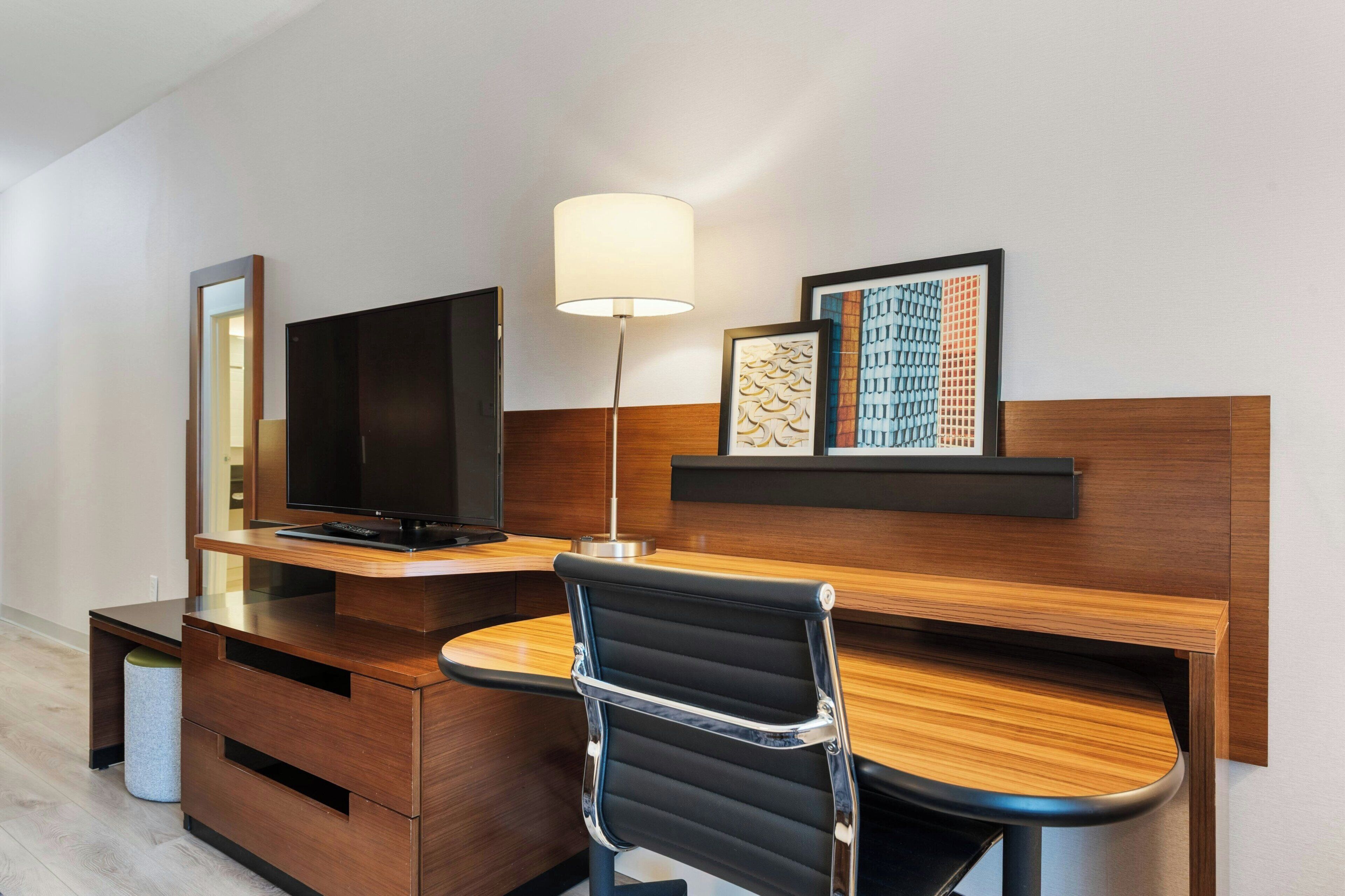 Una habitación de hotel moderna con un escritorio de madera, una butaca de cuero negro, un televisor de pantalla plana, una lámpara y obras de arte enmarcadas en la pared.