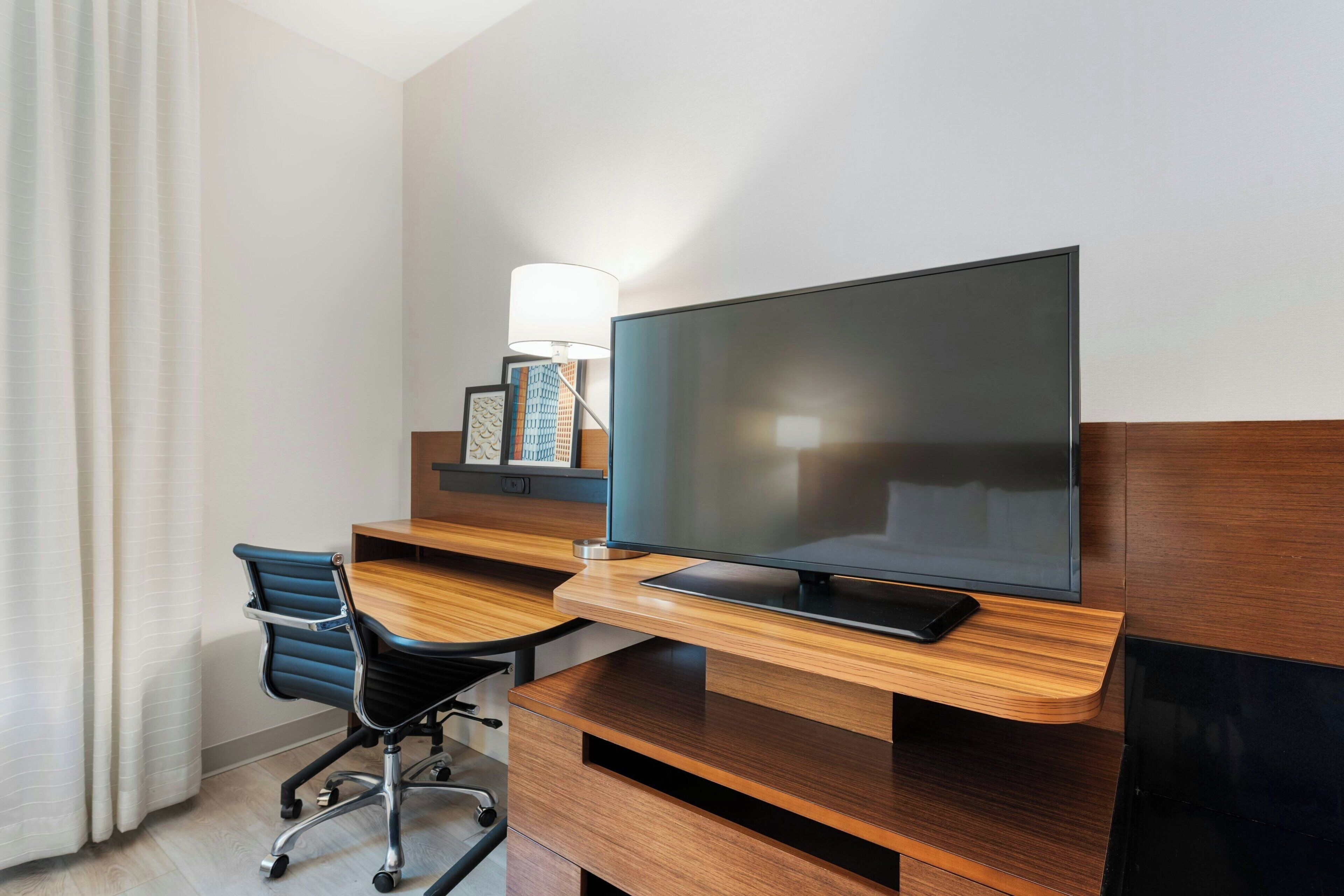 A hotel room with a wooden desk, a television, a chair, and a lamp.
